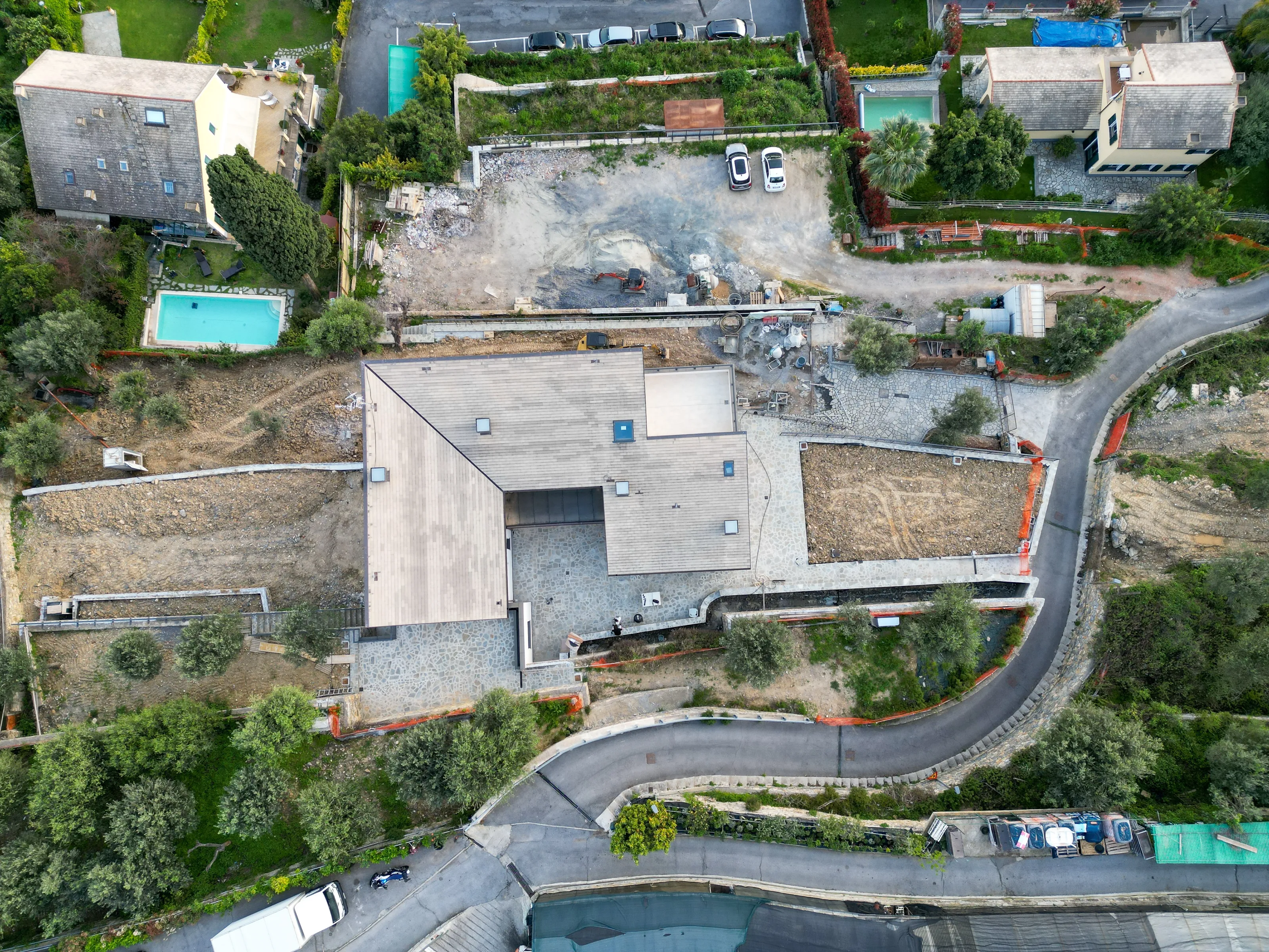 Top-down drone view of the house's roof among terraces of bare earth, a dirt yard with machinery and parked cars, and a road curving round the plot