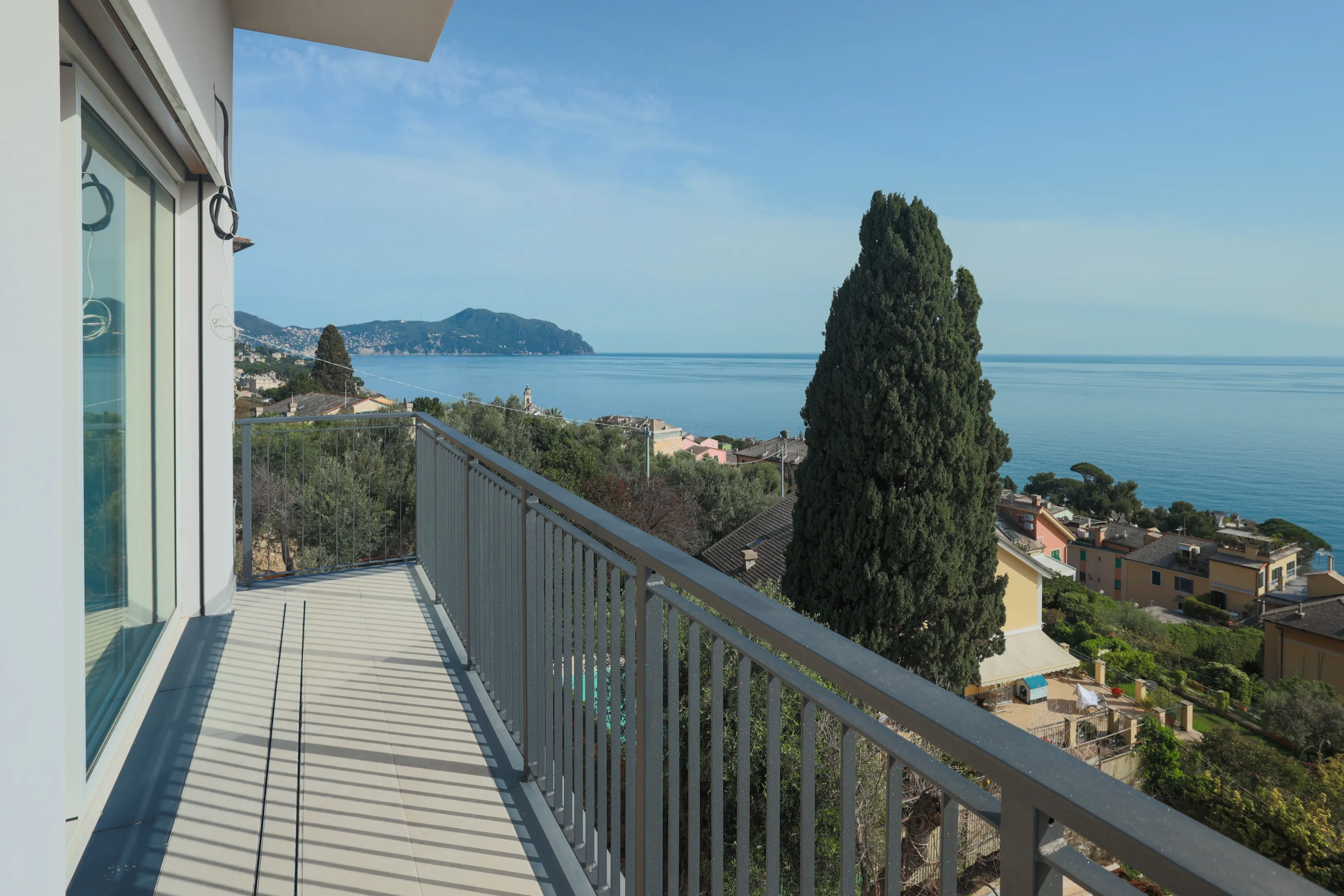 Un balcon au garde-corps gris le long de la maison, un grand cyprès, des toits en contrebas et la mer avec un promontoire au loin