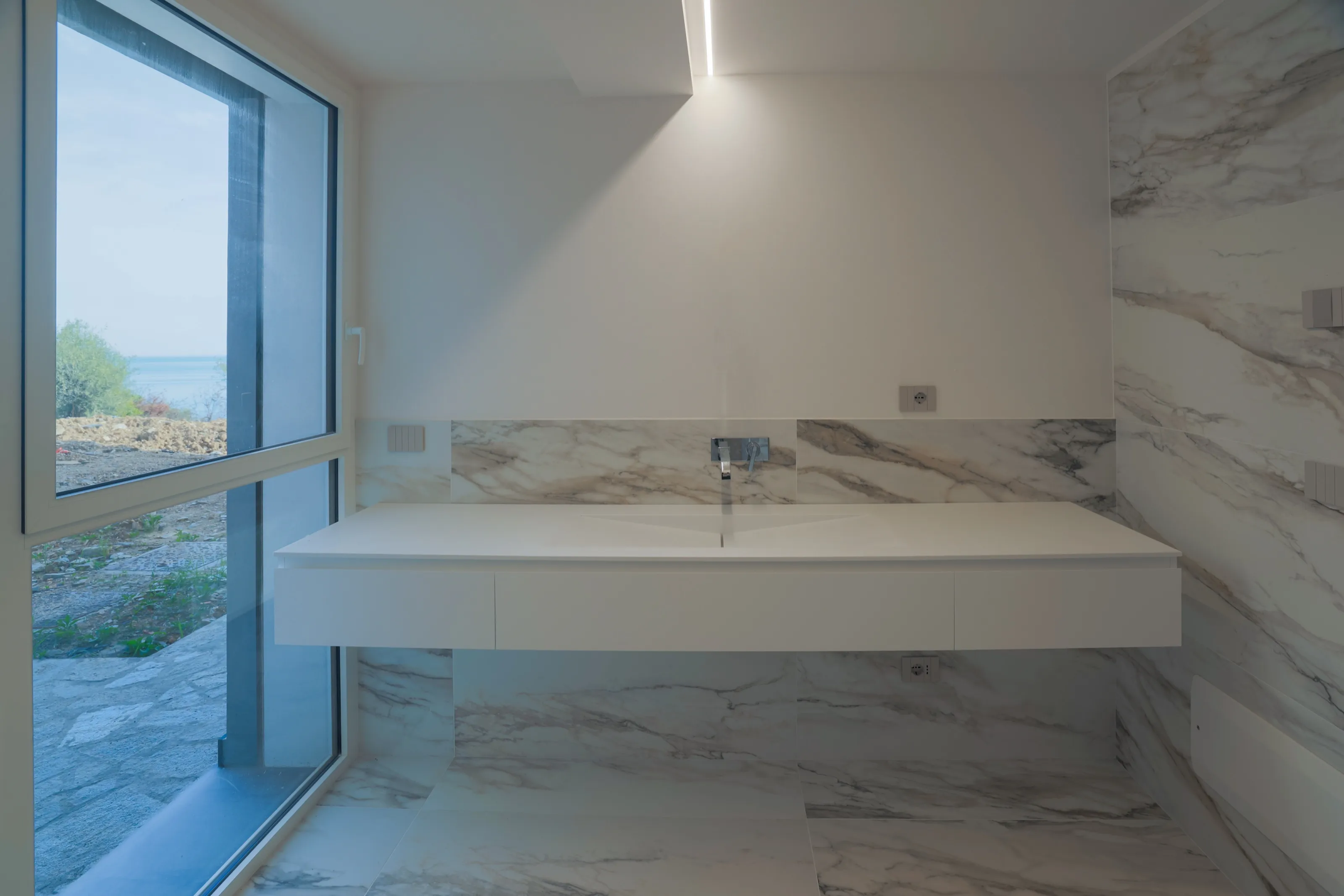 A white marble bathroom with a long floating basin and a tall window, the sea in the distance