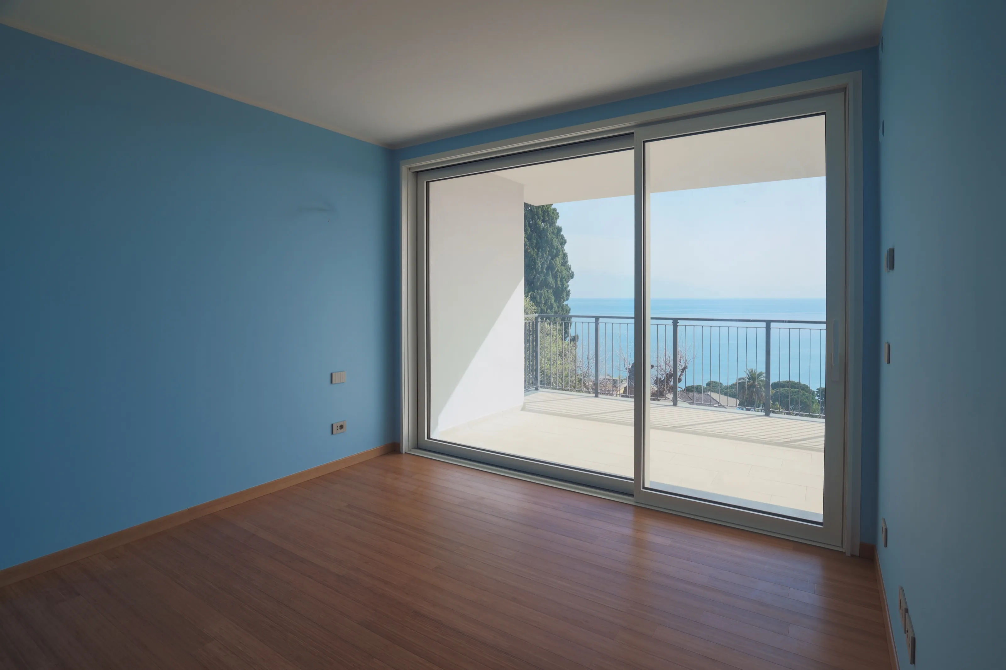 A bedroom with blue walls and a wooden floor, its sliding glass door giving onto a terrace with a railing and the sea beyond