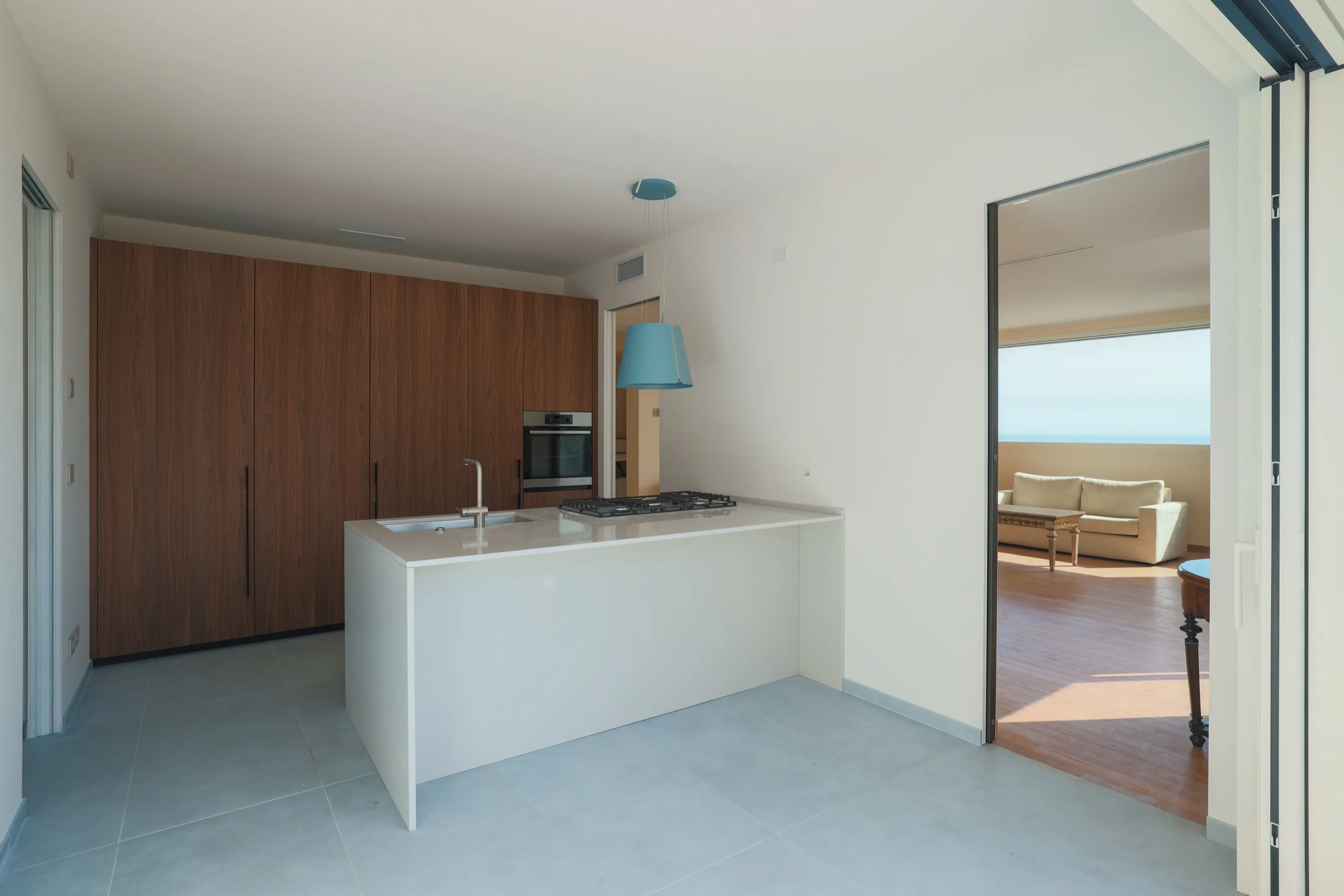 A kitchen with a white island, a pale blue pendant lamp and a wall of wood cabinets, opening onto a living room with a window on the sea