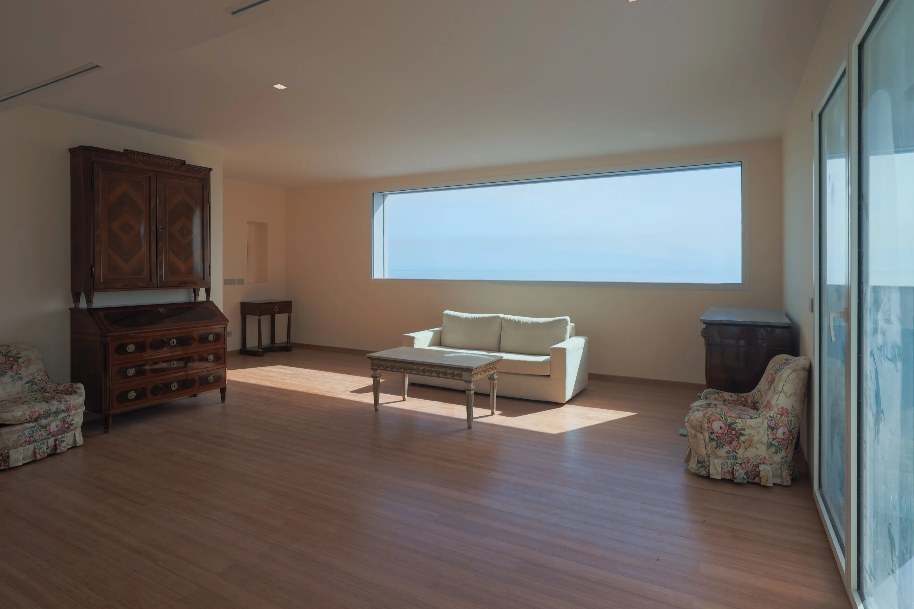 A living room with a long panoramic window on the sea, a white sofa, flowered armchairs and antique wooden furniture