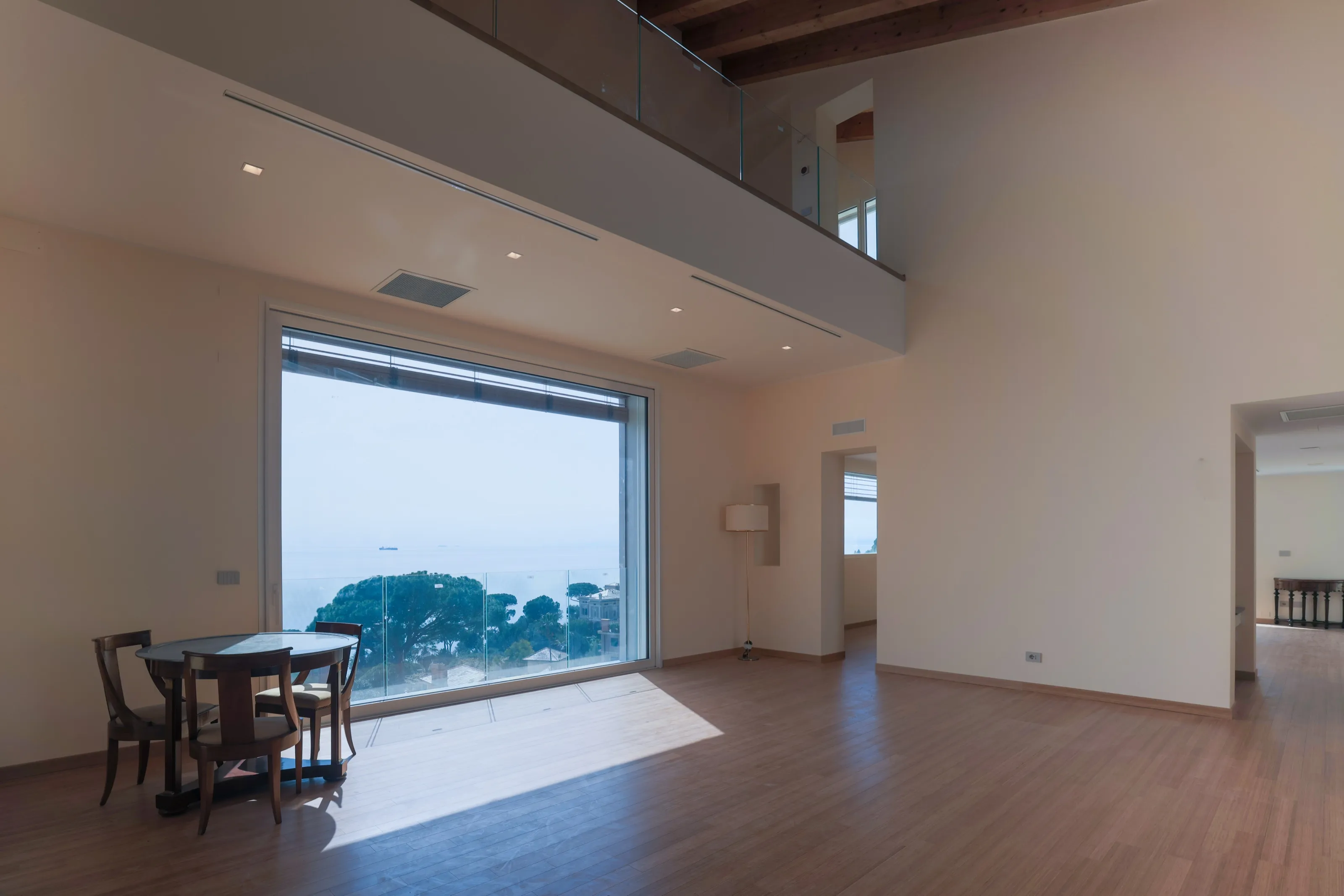 A double-height living room with a gallery above, a wooden floor, a small round table with chairs and a tall window framing pines and the sea