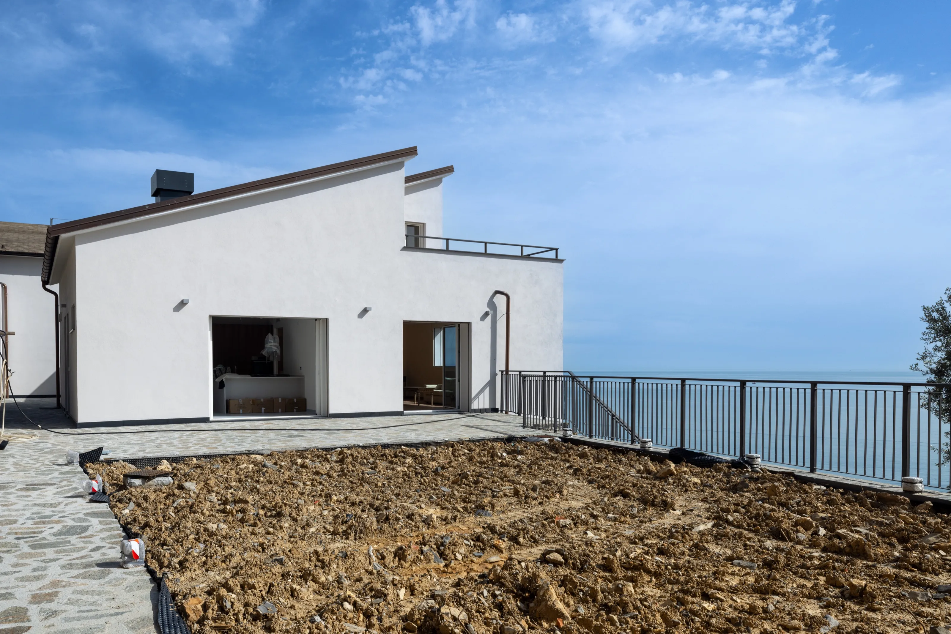 A white house with sloping roofs and a stone-paved terrace, a rectangle of bare earth in front, a railing and the sea beyond