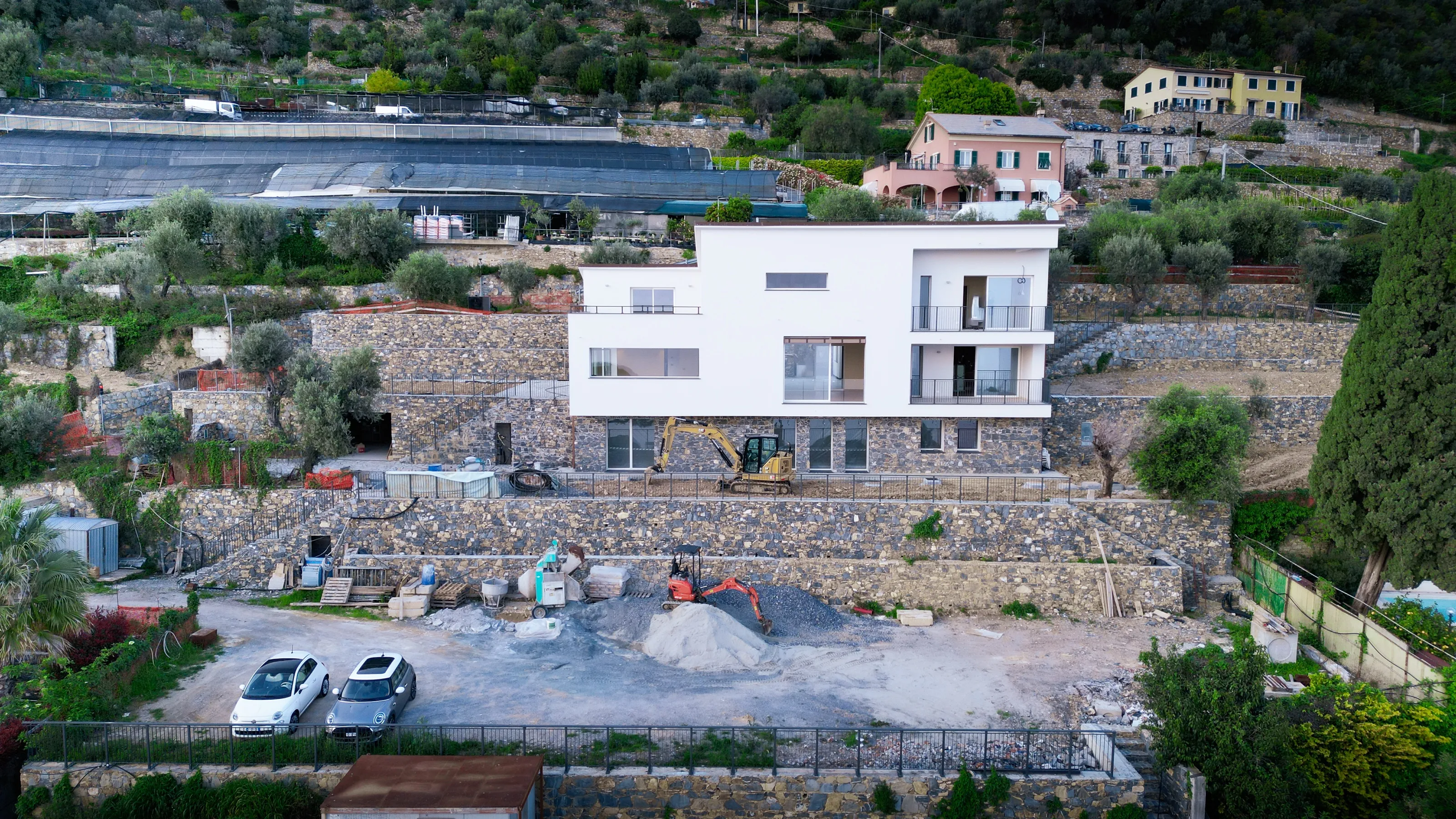 Drone photograph of a white three-storey house on stone terraces, with bare earth, a pile of gravel, two diggers and two parked cars in the yard below it
