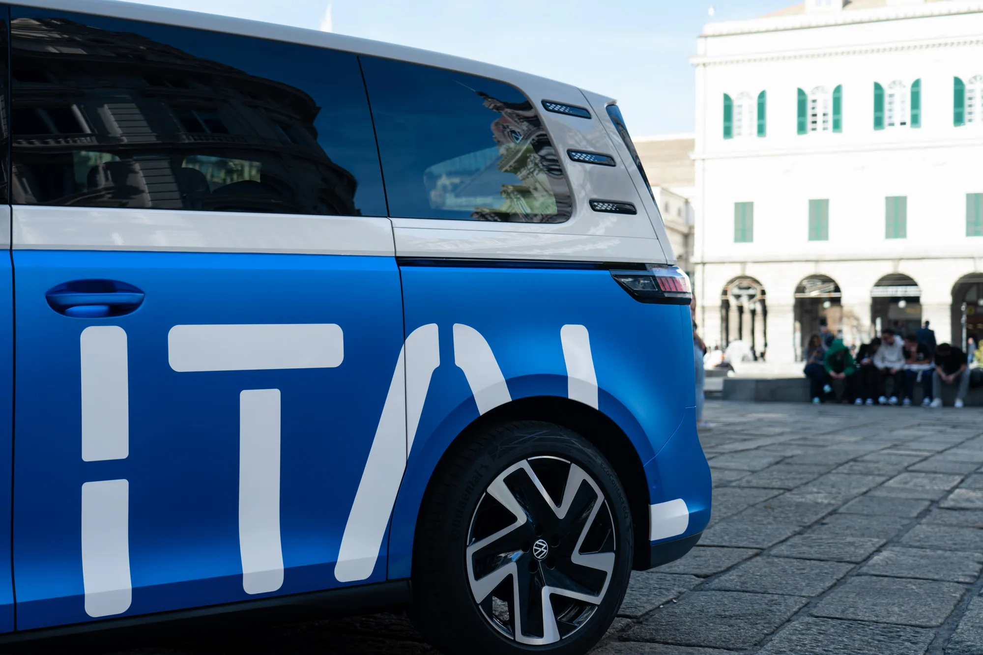 The rear half of the ID. Buzz, with large white ITAL letters on its blue side, in a Genoa square with people sitting in the background