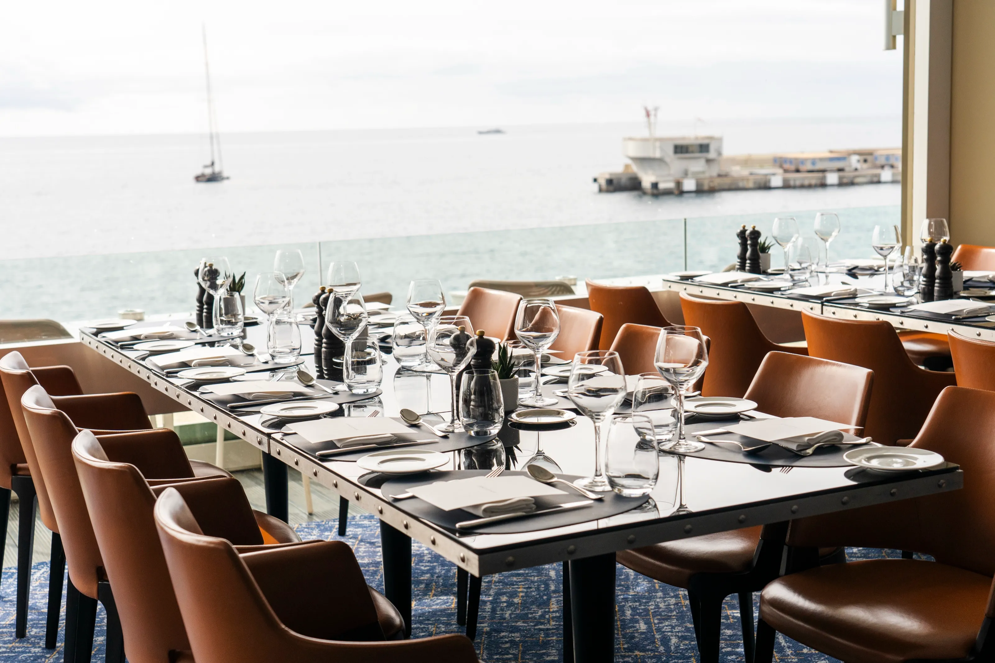 Long tables laid for lunch with glasses and tan leather chairs, beside a glass railing over the sea, a sailing boat on the horizon