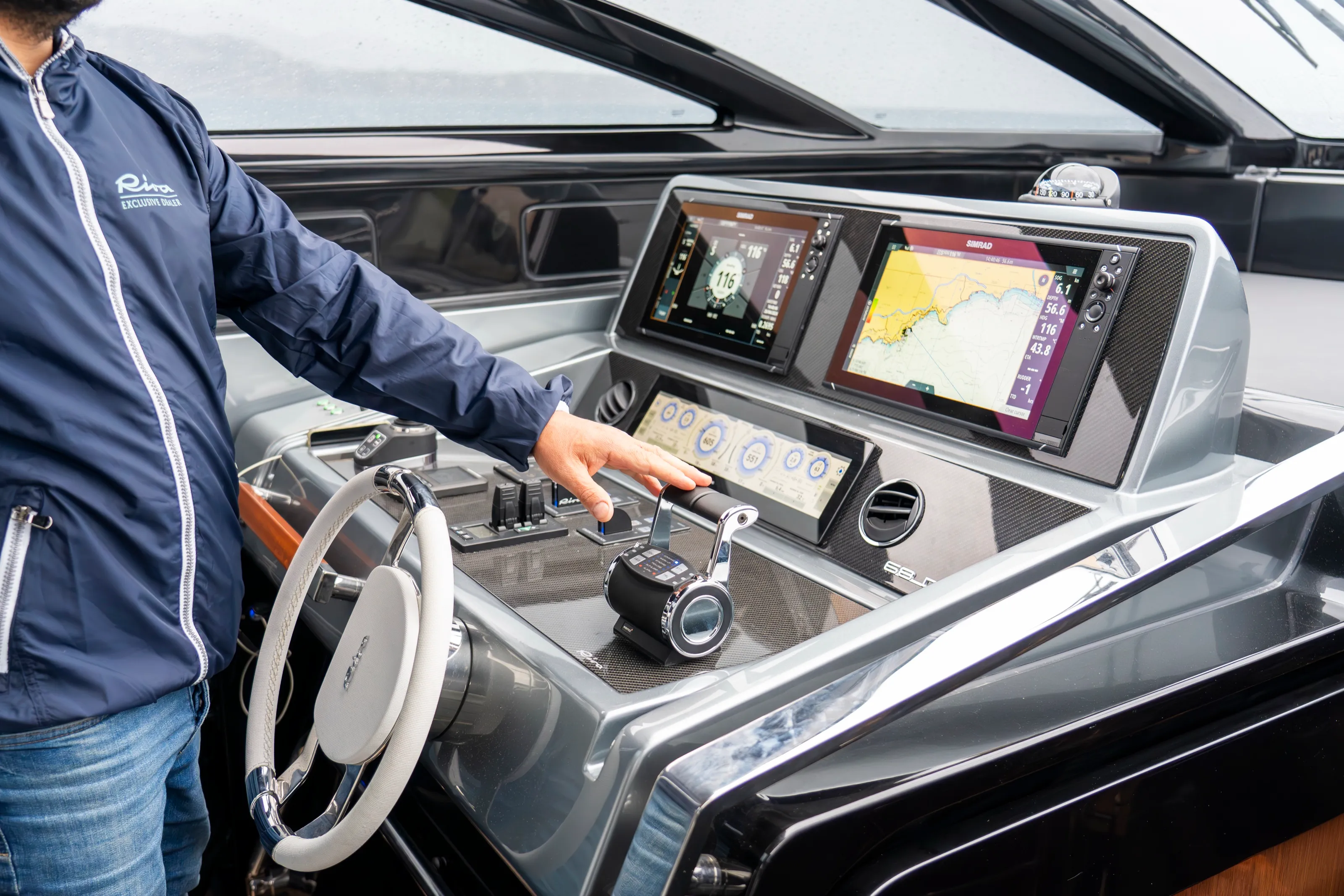 The helm of the Riva, a man in a navy jacket with his hand on the throttle, and two navigation screens, one showing a chart of the coast