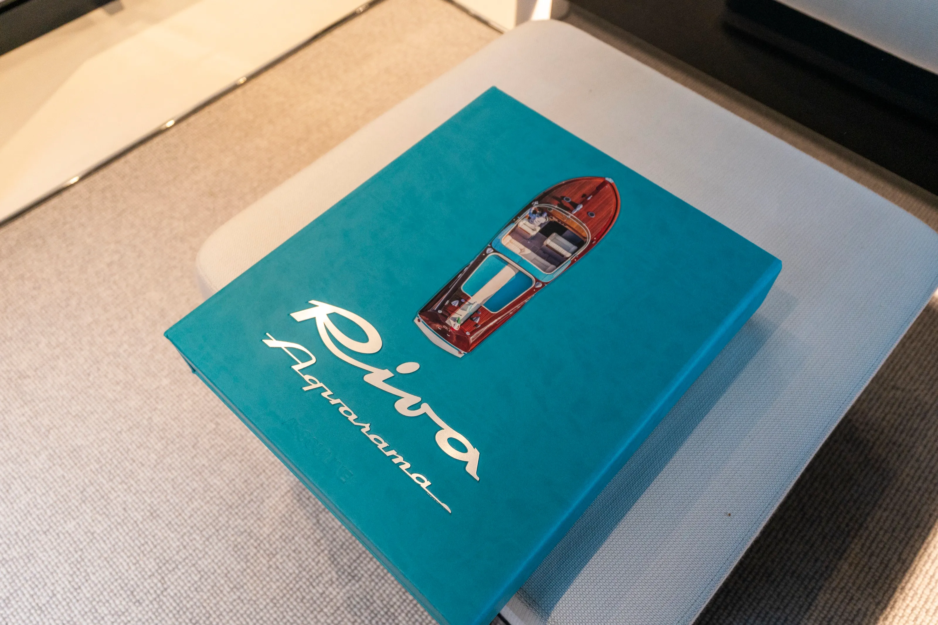 A teal Riva Aquarama book, a wooden boat seen from above on its cover, resting on a grey cushioned seat