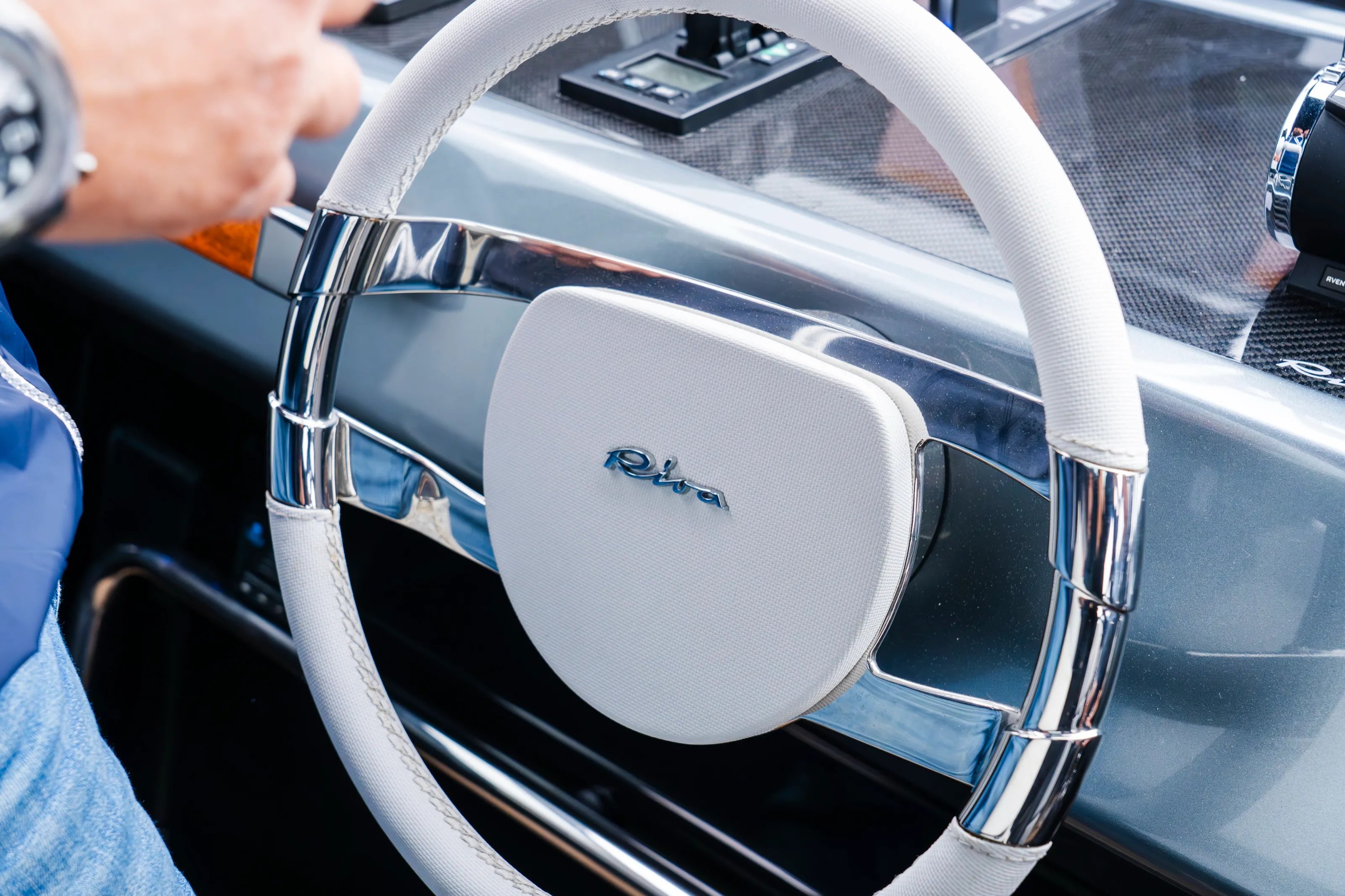 A hand on the white steering wheel of a Riva boat, the Riva script at its centre