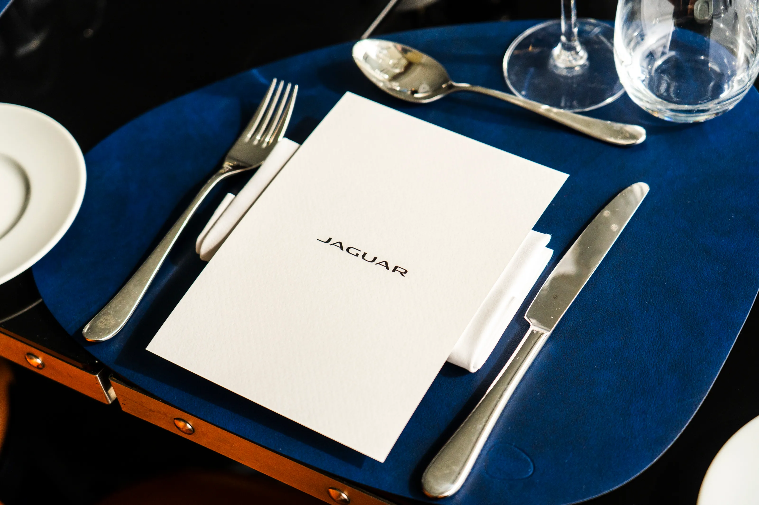 A place setting on a navy table mat, a white Jaguar menu between the fork and the knife, glasses behind