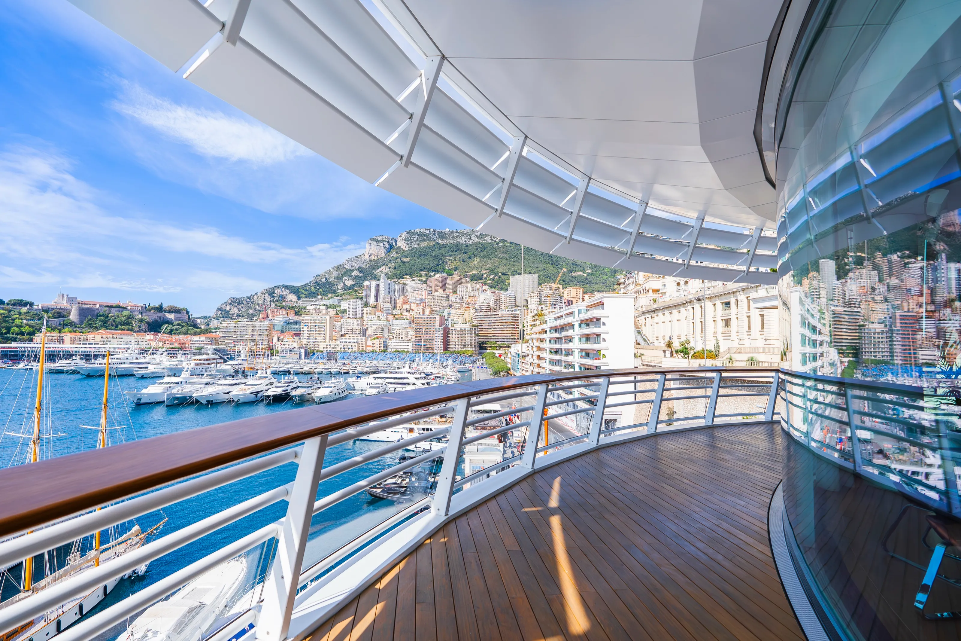 A curved hospitality terrace with a timber deck and glass railing under a white canopy, looking over Port Hercule and the city