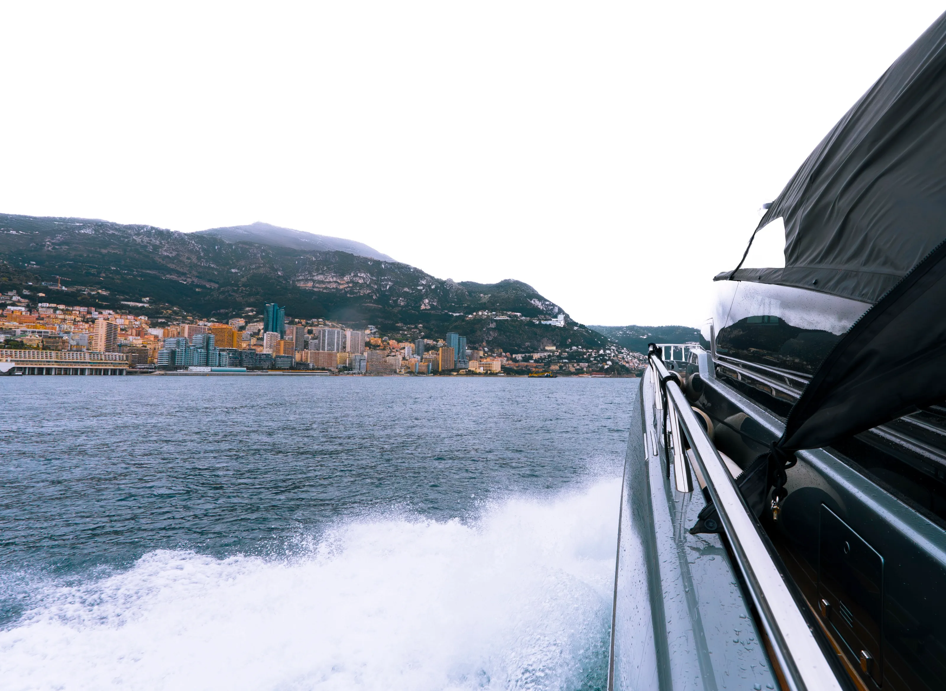 The boat's white wake on grey water, the buildings of Monaco and the mountains behind, the grey hull and its rail on the right