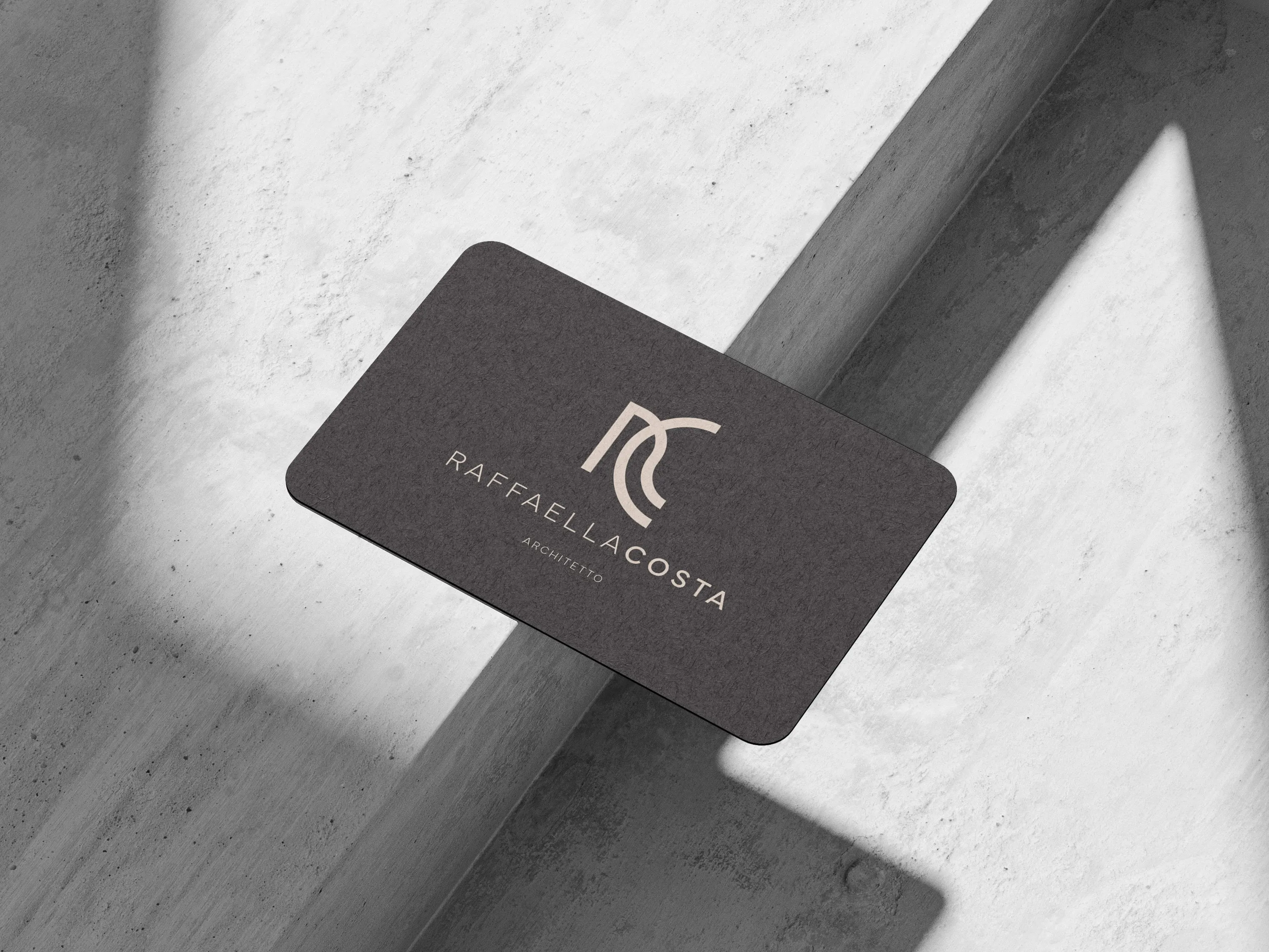 Mockup of a charcoal business card with rounded corners, the RC monogram, RAFFAELLA COSTA and ARCHITETTO printed in sand colour, lying on concrete striped with light and shadow