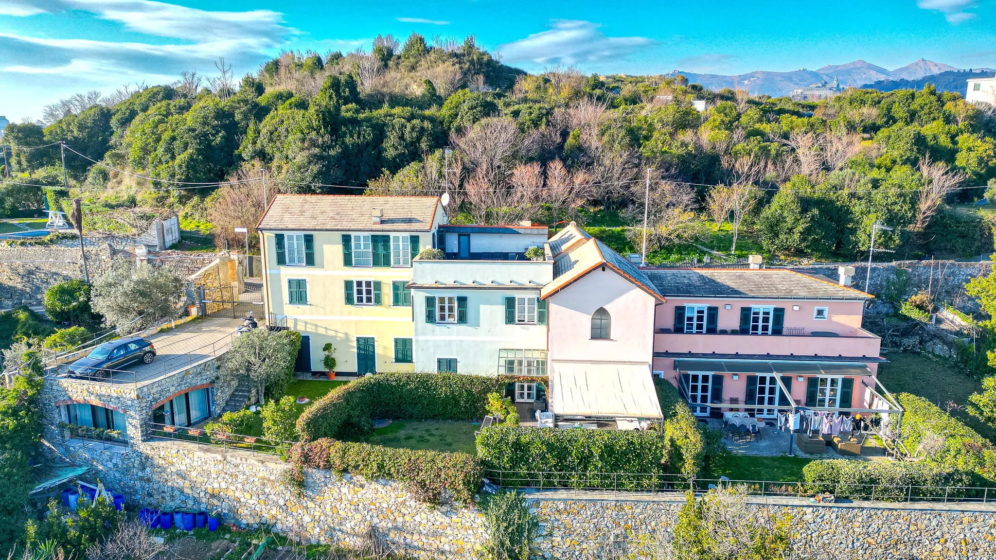 Casa Bianca seen from a drone: a row of joined houses painted yellow, pale green and pink on a wooded hillside, with gardens and a stone retaining wall in front, and mountains under a blue sky behind