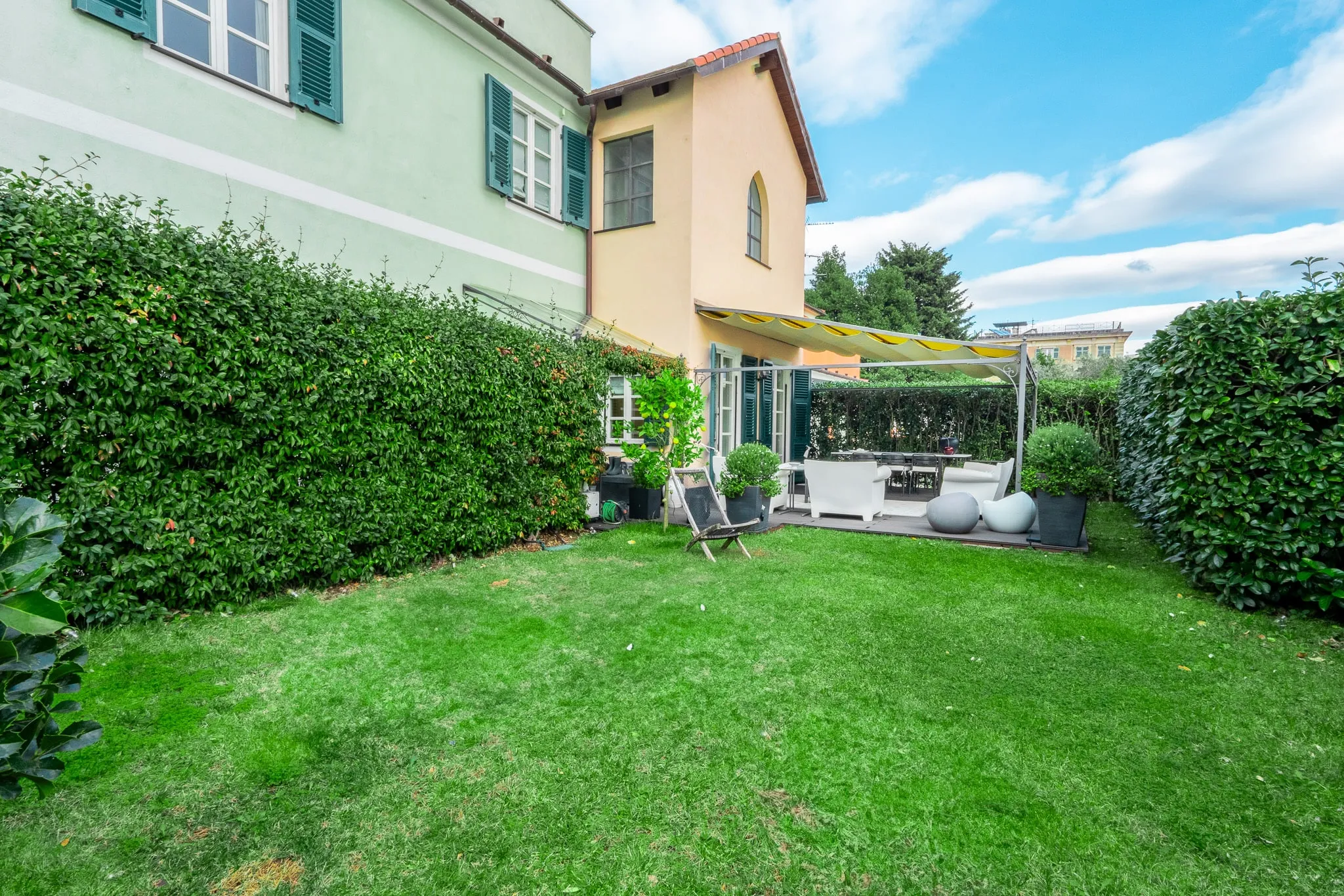 A lawn between tall hedges in front of a pale green house with green shutters and a peach-coloured gabled wing, with white outdoor sofas under an awning and a blue sky with white clouds