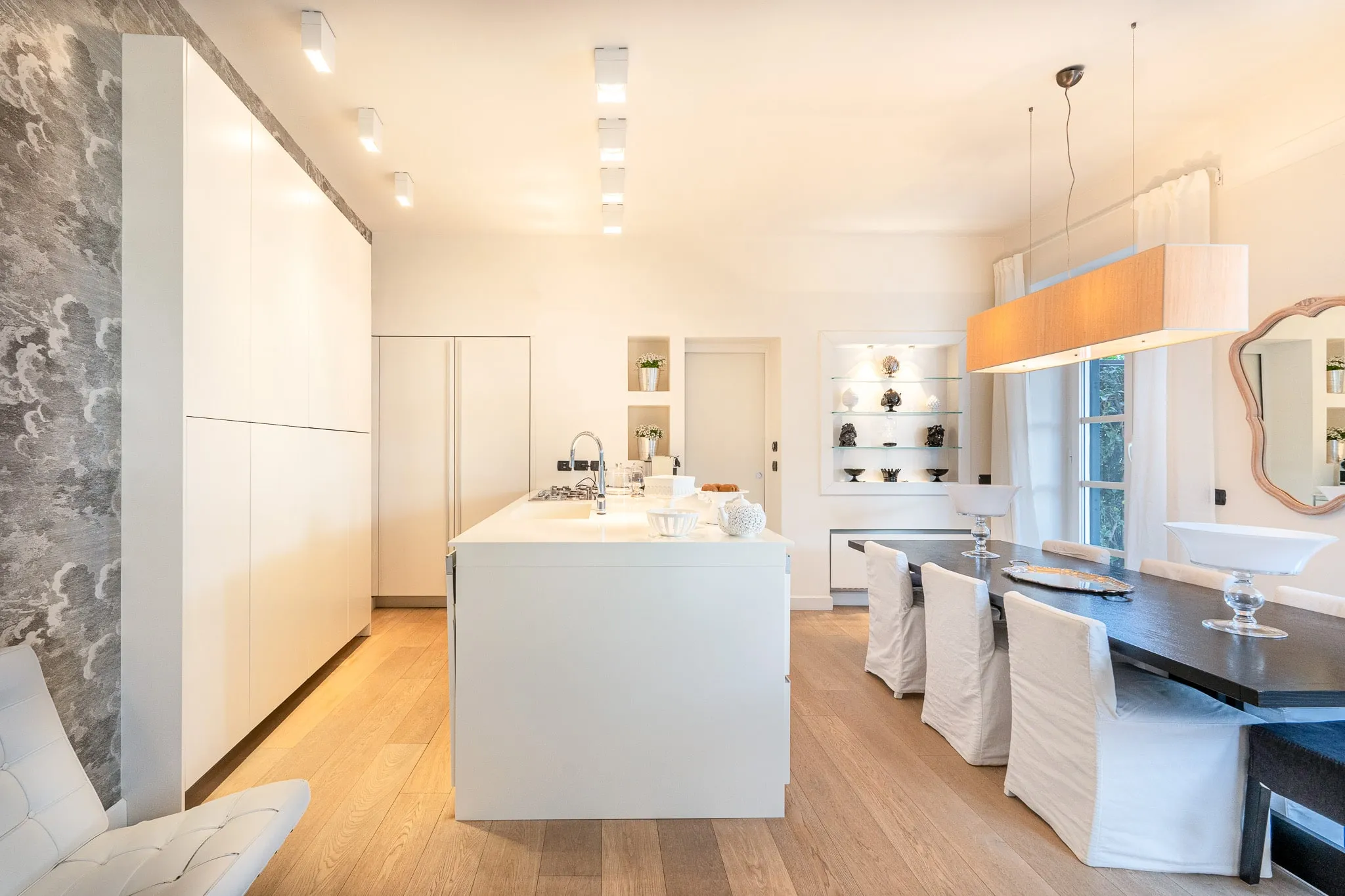 A white kitchen with a long island and tall cupboards on a pale wooden floor, and beside it, on the right, a long dark dining table with white-covered chairs under a long pendant lamp