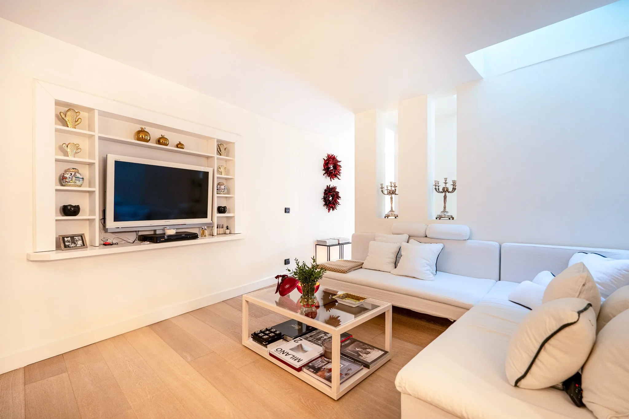 A white living room with a long L-shaped sofa and pale cushions, a glass coffee table stacked with books, a television set into white shelving, two candlesticks in high niches and a skylight above