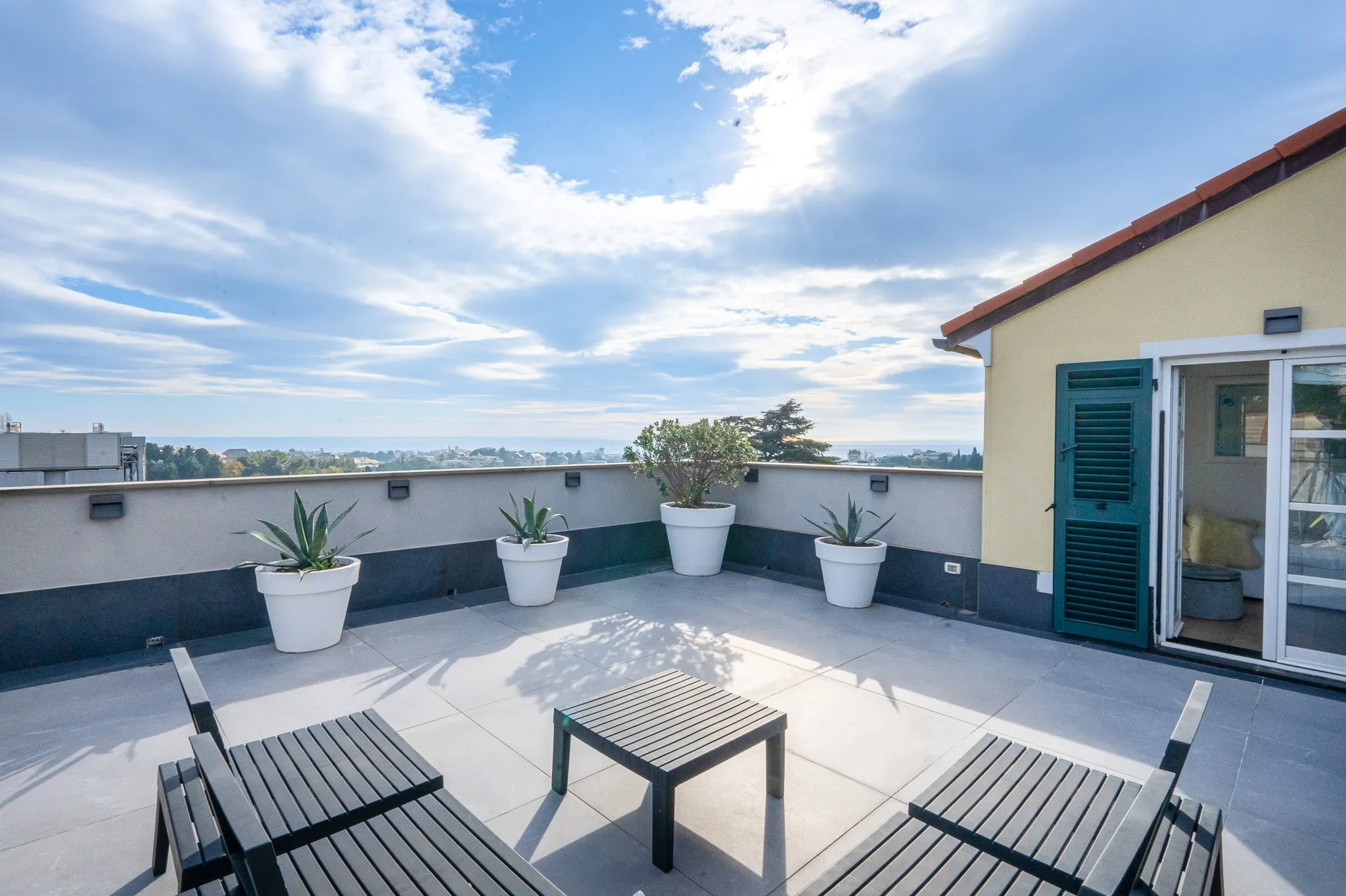 A roof terrace with two dark wooden loungers, a low table and potted agaves along the parapet, beside a yellow wall with a green shutter and open glass doors, looking out over trees and rooftops to the sea