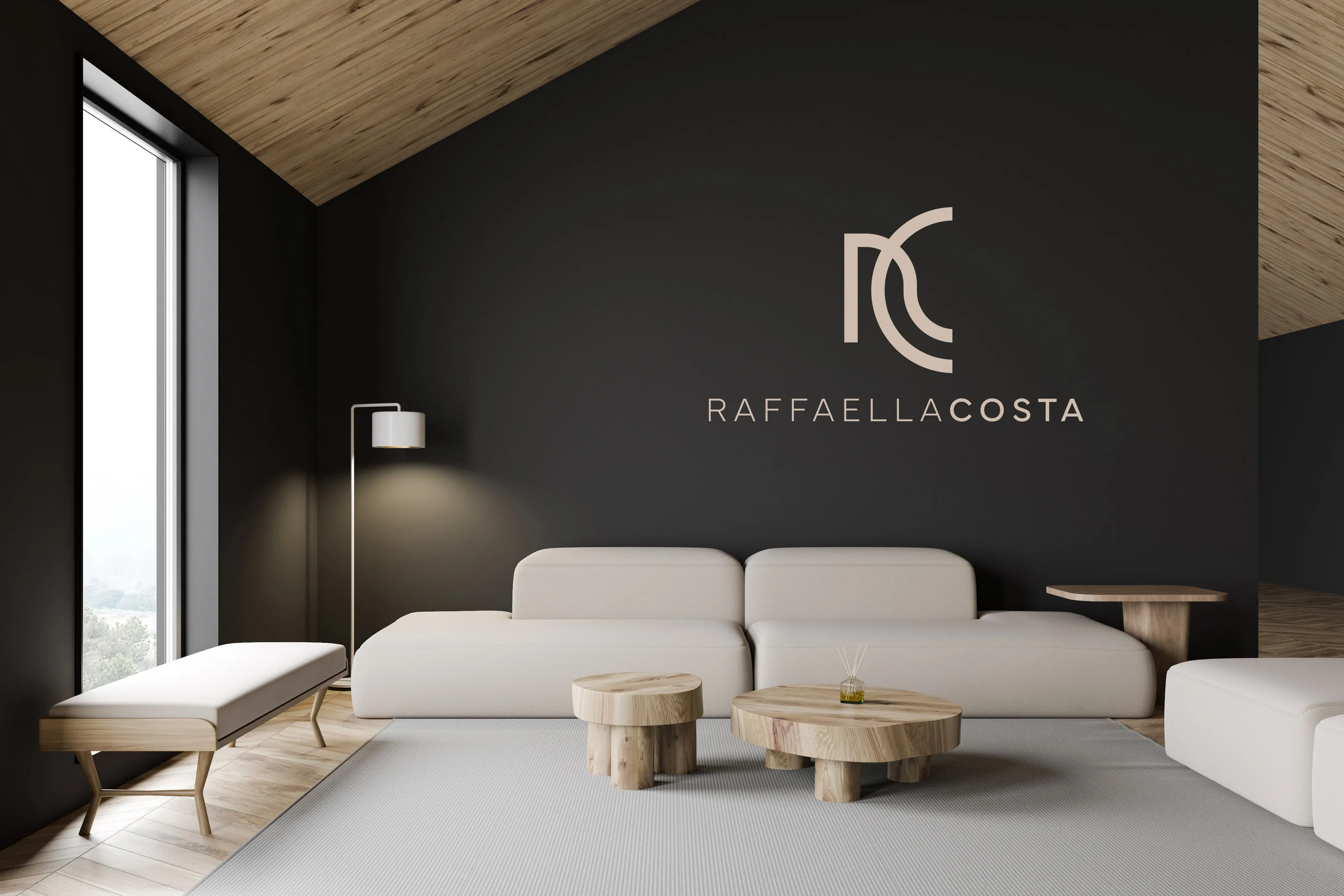 Mockup of the Raffaella Costa monogram and wordmark in sand-coloured letters on a charcoal wall, above a long pale sofa and two round wooden tables, under a sloping timber ceiling