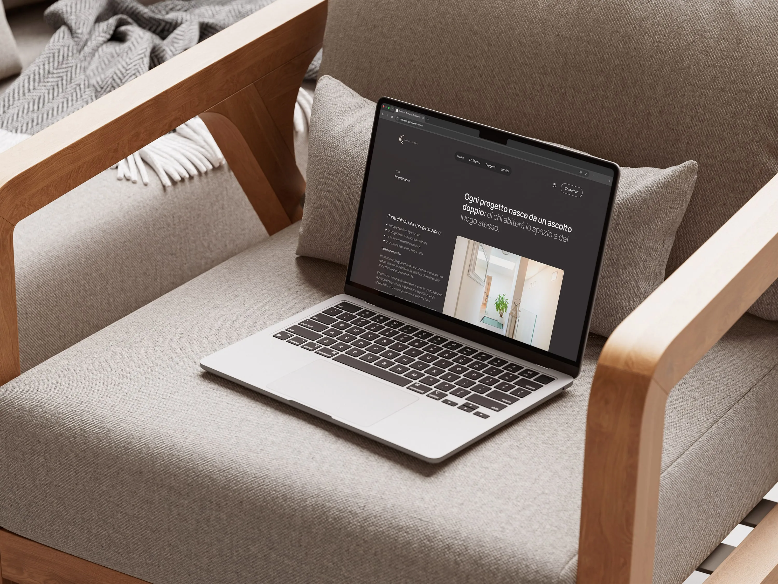 Mockup of a laptop on a fabric armchair with wooden arms, open on the Servizi page of raffaellacosta.com: the Progettazione section, a heading in Italian, a list of key points and a photograph of a white interior with a plant