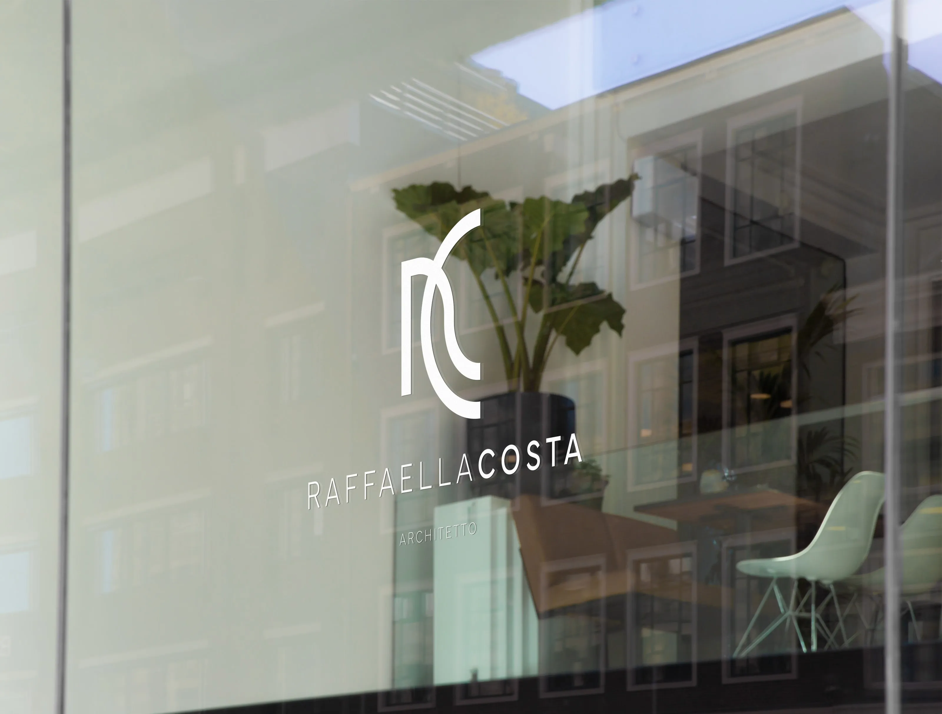 Mockup of the monogram, the wordmark and ARCHITETTO in white on a shop window that reflects the buildings across the street, with a large-leaved plant and pale chairs inside
