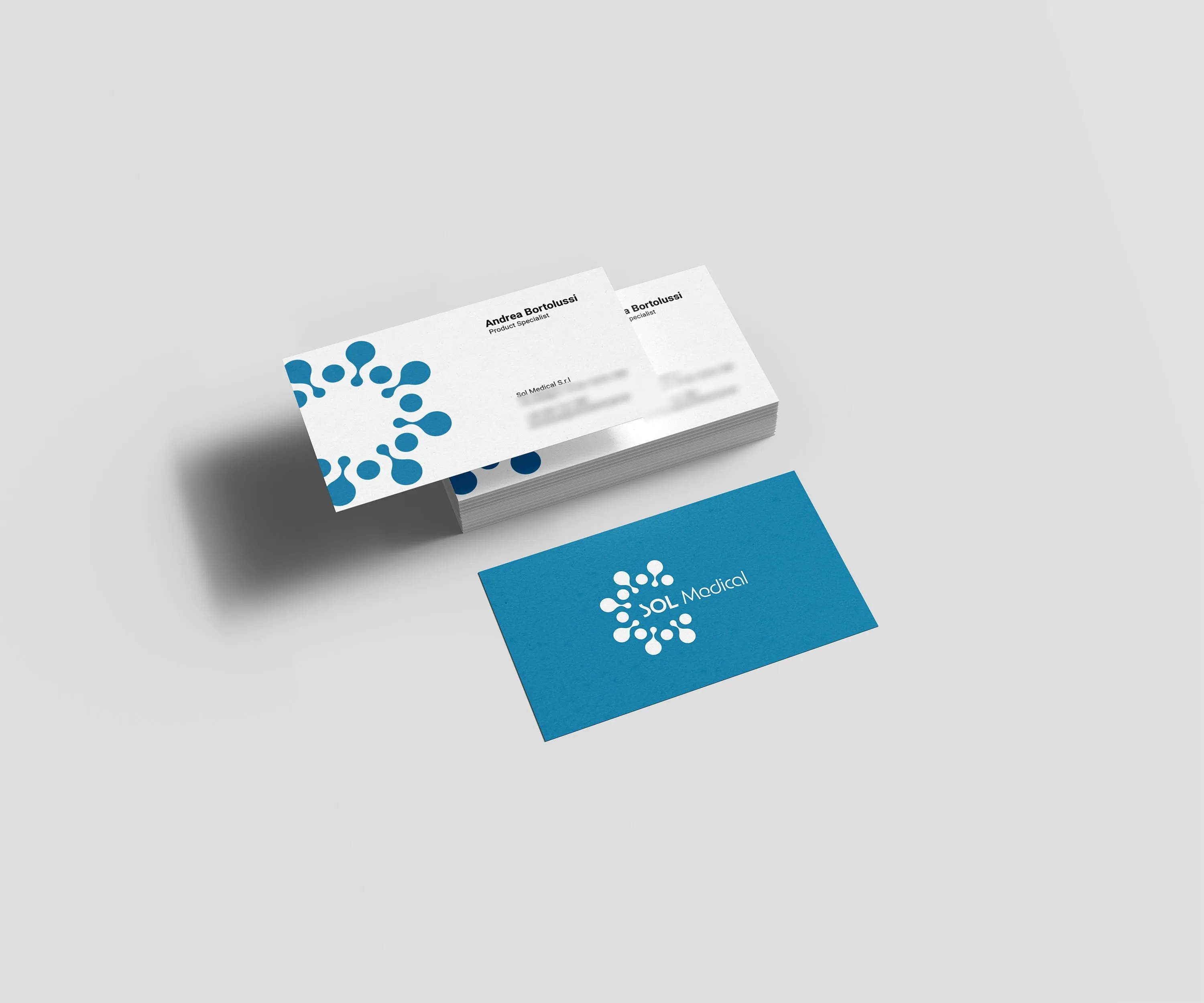 Mockup of SOL Medical business cards on a pale grey surface: a stack of white cards with a large cerulean icon running off the left edge and blurred contact lines, and one cerulean card with the full logo in white